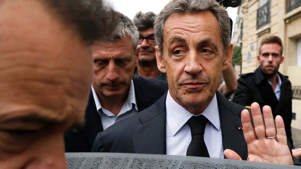 Nicola Sarkozy umgeben von Männern grüsst mit der offenen Handlfäche Journalisten