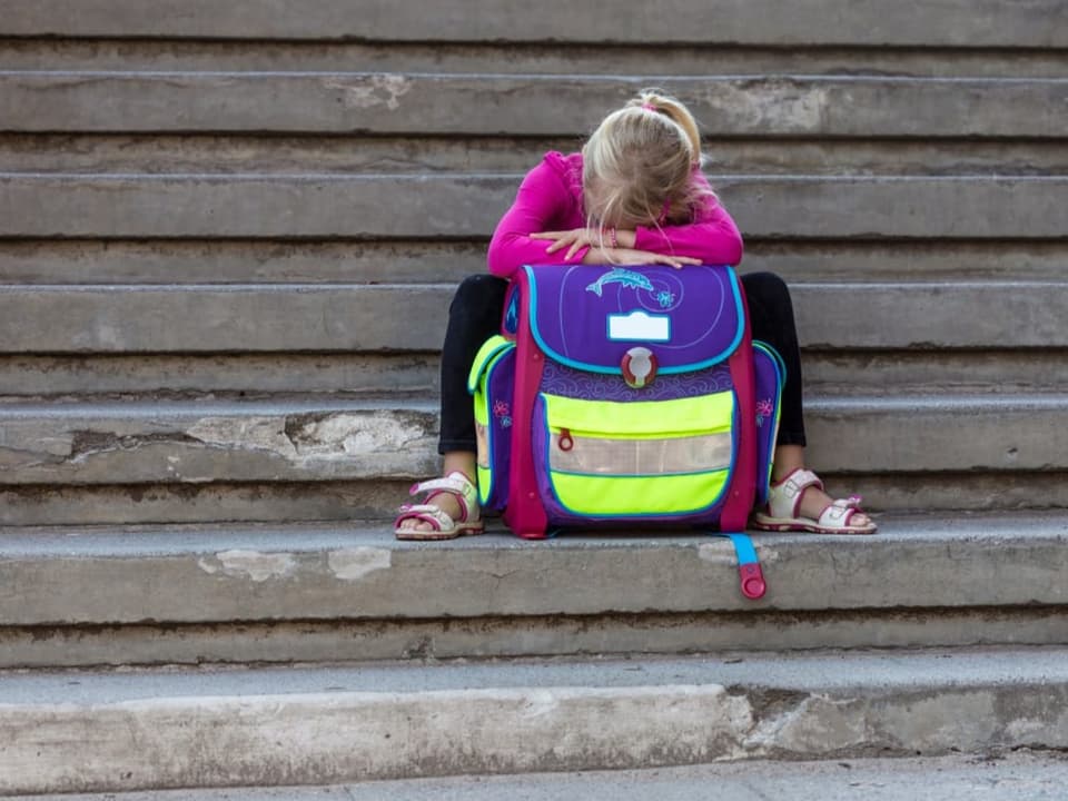 Ein kleines Mädchen sitzt traurig auf einer Treppe, das Gesicht in seinen Rucksack vergraben