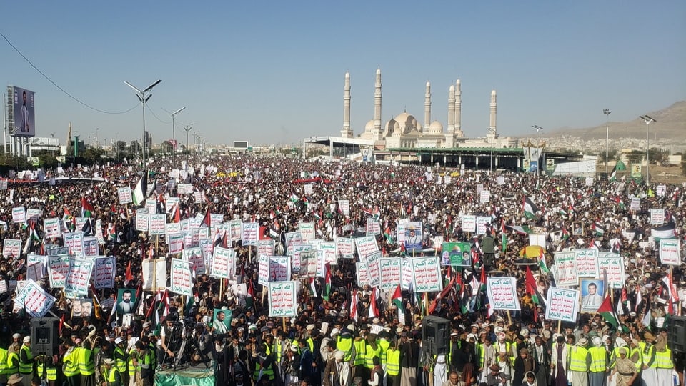 eine Menschenmenge mit arabisch beschrifteten Plakaten, im Hintergrund Minarette in Sanaa, Jemen