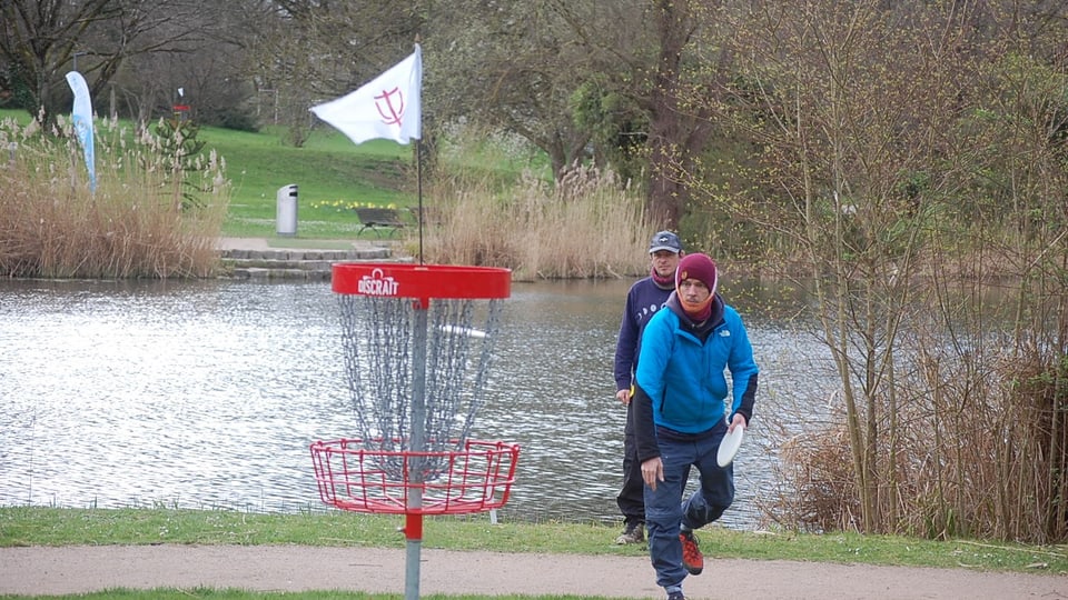 Zwei Personen spielen Discgolf an einem Teich im Park.