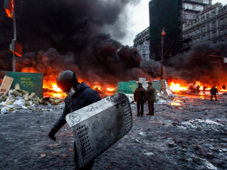 Brennende Barrikaden im Hintergrund, im Vordergrund ein vermummter Demonstrant mit einem von einem Polizisten erbeuteten Schutzschild.