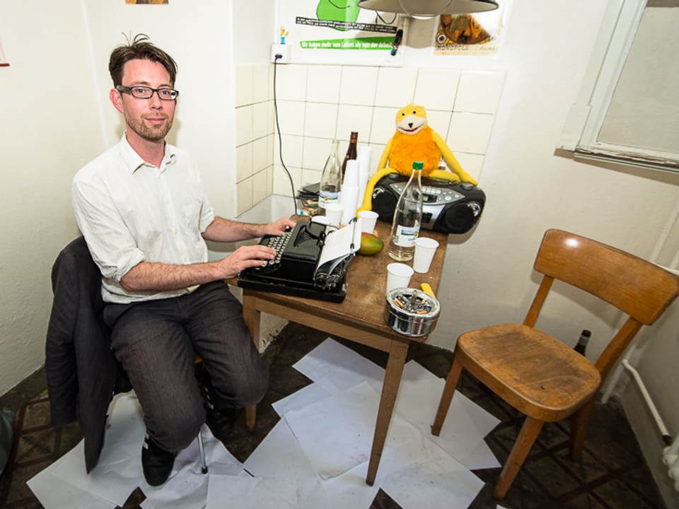Ein Mann sitzt in einer Küche vor einer Schreibmaschine. Am Boden liegen viele Papierblätter.
