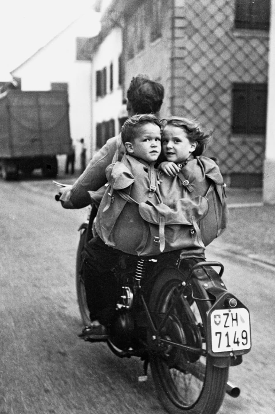 Schwarz-Weiss-Bild: Ein Mann auf einem Motorrad, auf dem Gepäckträger eine Tasche, in der ein Knabe und ein Mädchen sitzen.