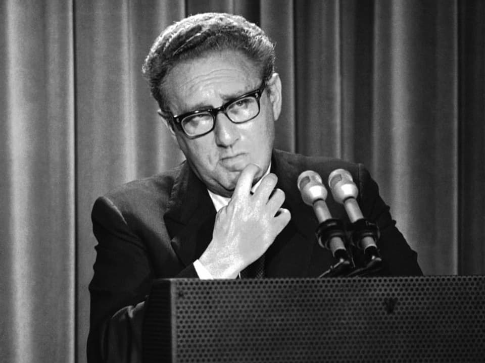 Henry Kissinger steht hinter einem Pult mit zwei Mikrofonen.
