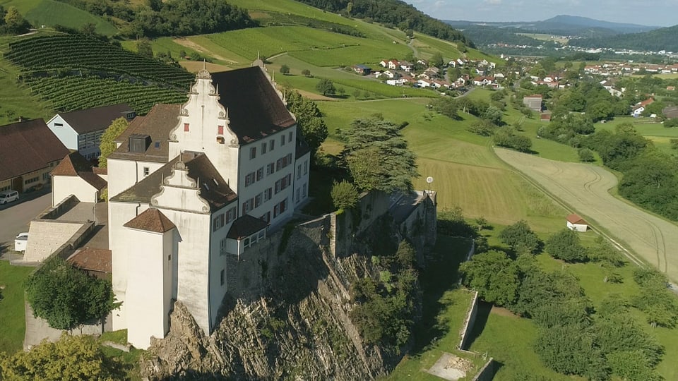 Luftaufnahme einer Schlossanlage auf einem Felsen, eingebettet in einer hügeligen Landschaft.