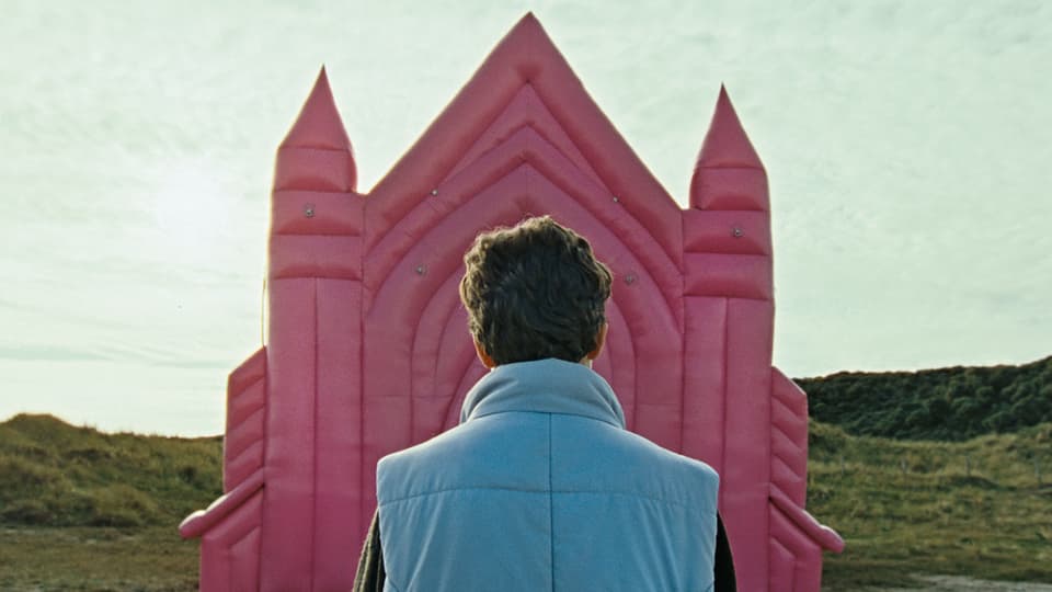 Eine kurzhaarige Person steht vor dem Eingang einer rosaroten Hüpfburg, der einer Vulva gleicht.