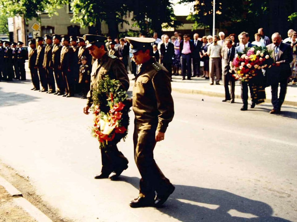 Zwei Soldaten in Uniform tragen eine Blumenkranz. Dahinter weitere Soldaten in einer Reihe.