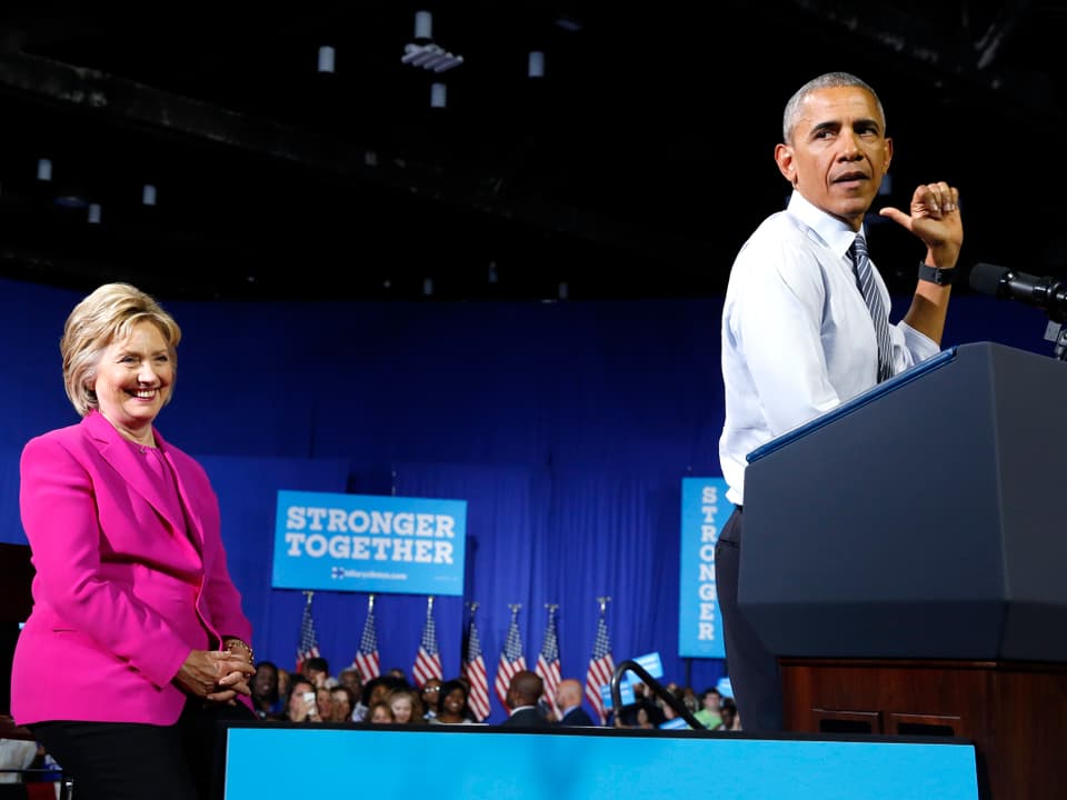Barack Obama an einem Rednerpult zeigt auf Hillary Clinton hinter sich.