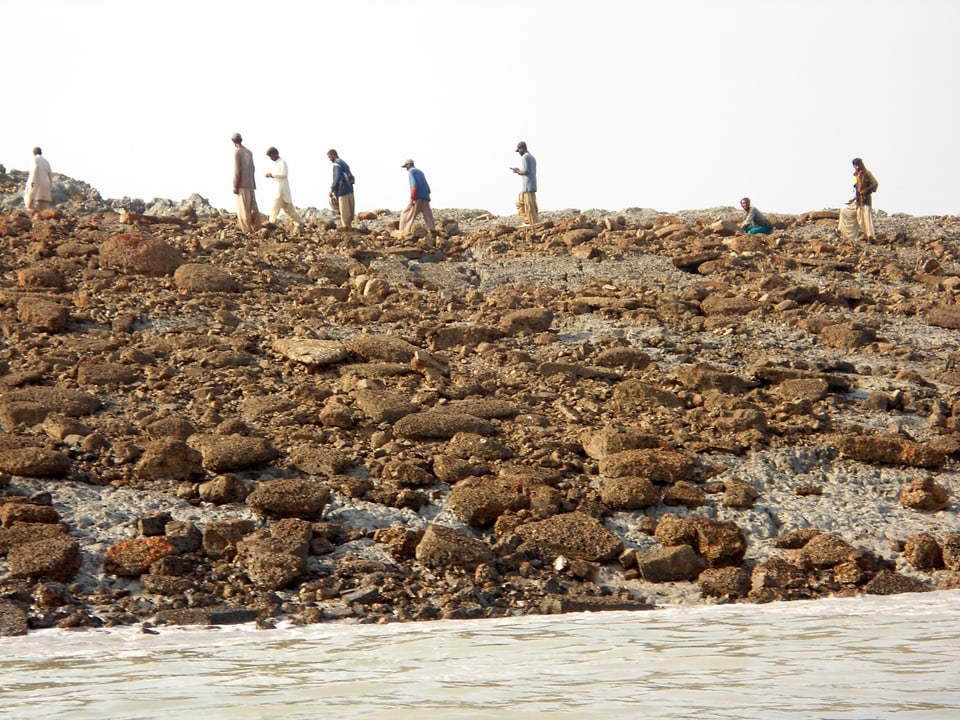 Vereinzelte Menschen laufen auf einer Insel, auf ihr liegen bräunliche Steine. 