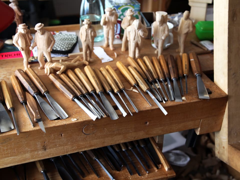 Viele verschiedengrosse Holzbearbeitungswerkzeuge liegen auf dem Tisch der Werkstatt. Im Hintergrund stehen, sich im Verarbeitungsprozess befindende, Holzfiguren.  