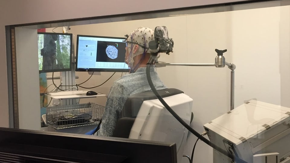 Mann in einem Forschungslabor. Er trägt eine Mütze, an der Kabel befestigt sind. Auf einem Bildschirm ist ein Hirn zu sehen.