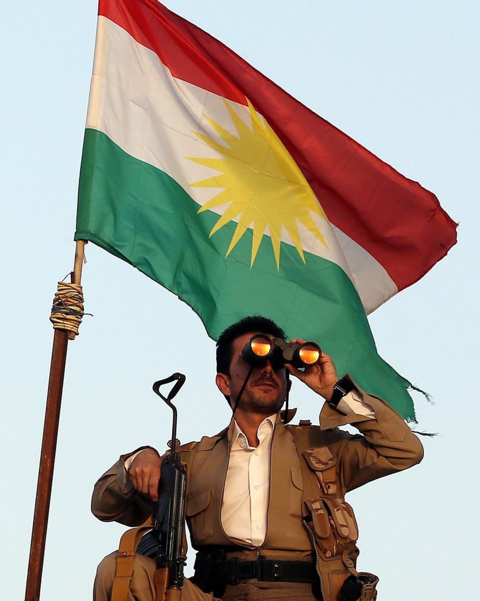 Ein Peschmerga-Kämpfer mit Sturmgewehr und Feldstecher vor einer Flagge Kurdistans.