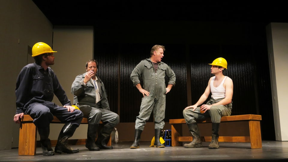 Szene in einem Landtheater: Arbeiter mit gelben Schutzhelmen stehen und sitzen auf der Bühne. 