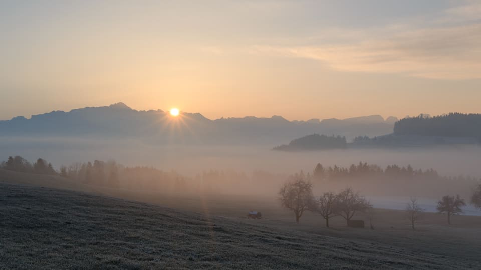 Die Sonne geht hinter den Bergen auf, über dem Tal liegen flache Nebelfelder.