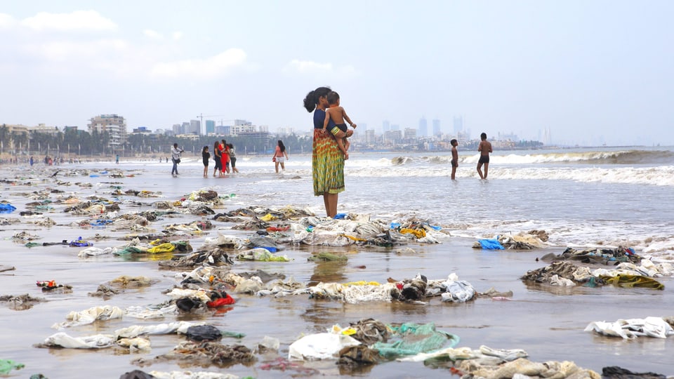 Eine Frau steht am Strand - viel Plastik wurde angespühlt, leere Flaschen, vor allem Plastiksäcke.