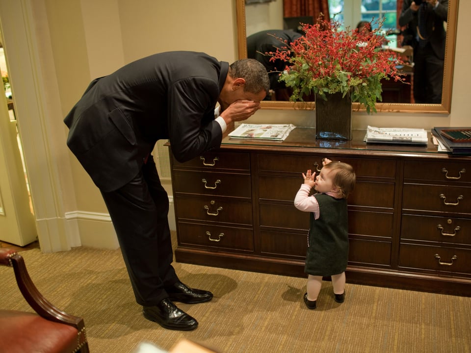 Obama hält sich die Hände vor das Gesicht und spielt mit einem kleinen Kind.