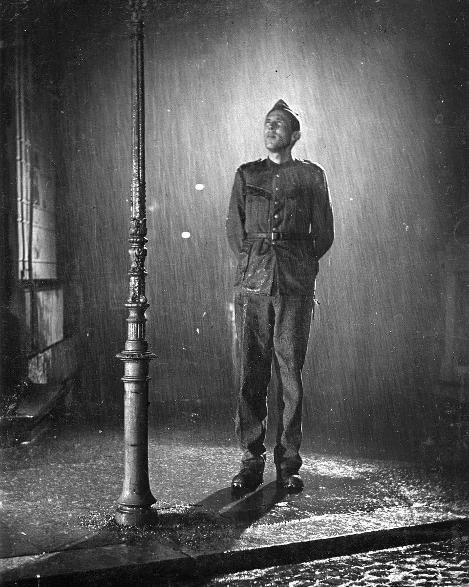 Ein junger Mann in Uniform steht bei ströhmendem Regen unter einer Strassenlaterne.