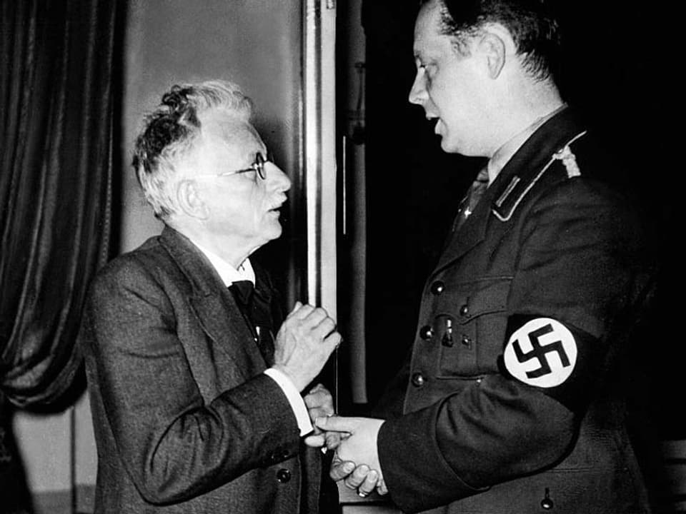 Ein Mann mit Schnauz unterhält sich mit einem Mann mit Swastika an der Uniform.