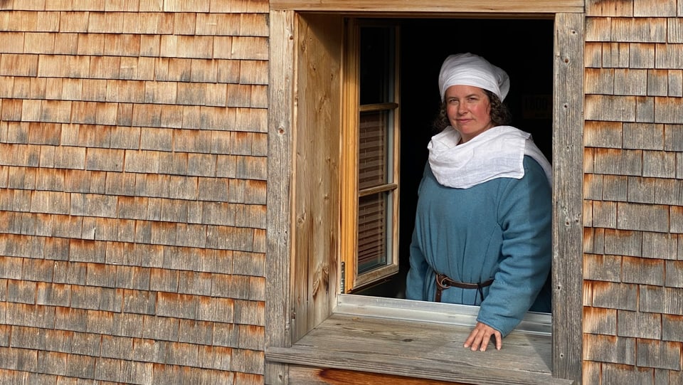 Ein Frau mittleren Alters, gekleidet wie im Hochmittelalter, schaut aus einem Holzhaus.