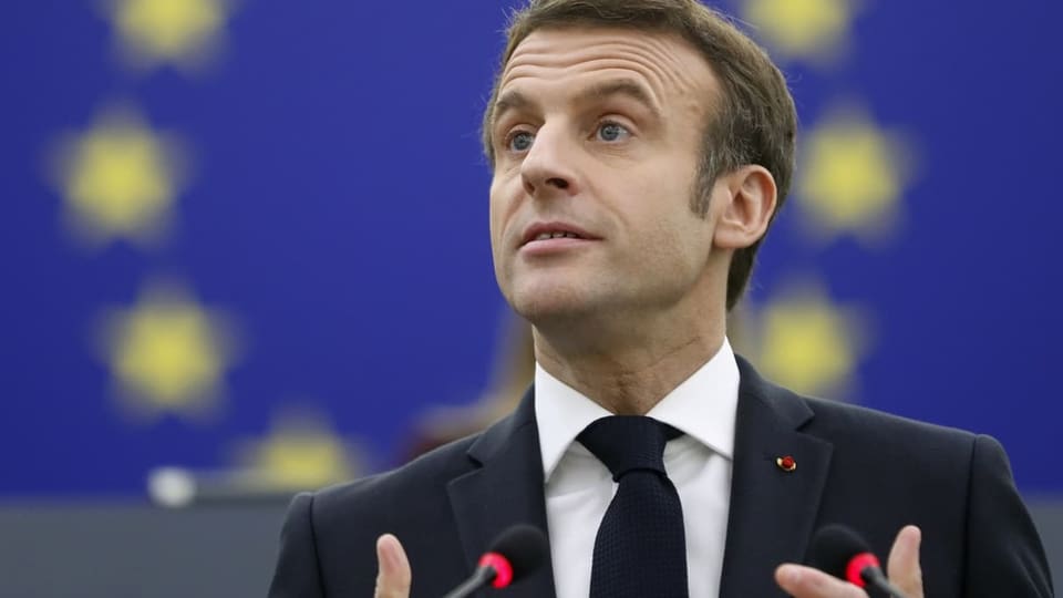 Macron gestikuliert beim Reden mit den Händen. Hinter ihm ist die blaue EU-Flagge zu sehen. 