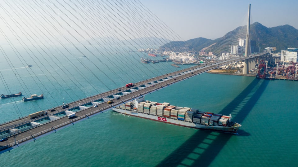 Ein Boot läuft in den Containerhafen von Hong Kong ein. Darüber verläuft eine Brücke. 