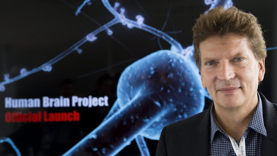 Henry Markram vor einem Plakat, das eine Synapse zeigt. Darauf steht geschrieben "Human Brain Project. Official Launch".