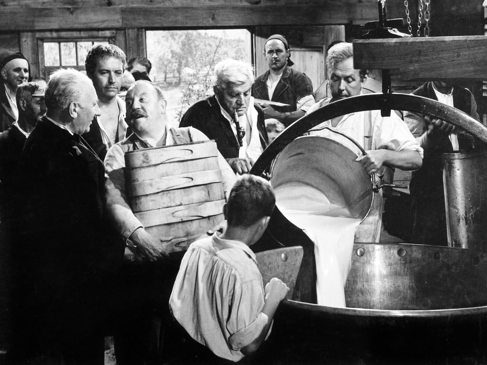 Szene in einer Molkerei. Mehrere Männer liefern Milch. Ein Kessel Milch wird gerade in einen grossen Bottich geschüttet. Ein Junge schaut dabei zu.