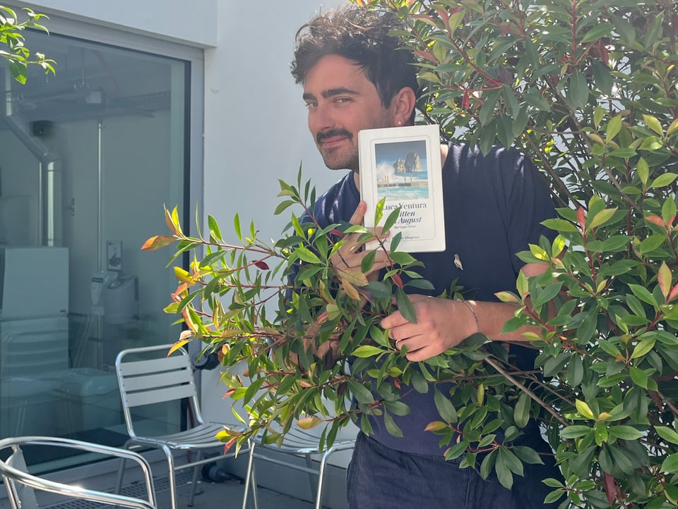 Moderator Dario Cantieni steht hinter einem Busch und präsentiert sein Lieblingsbuch mit einem verschmitzten Blick.