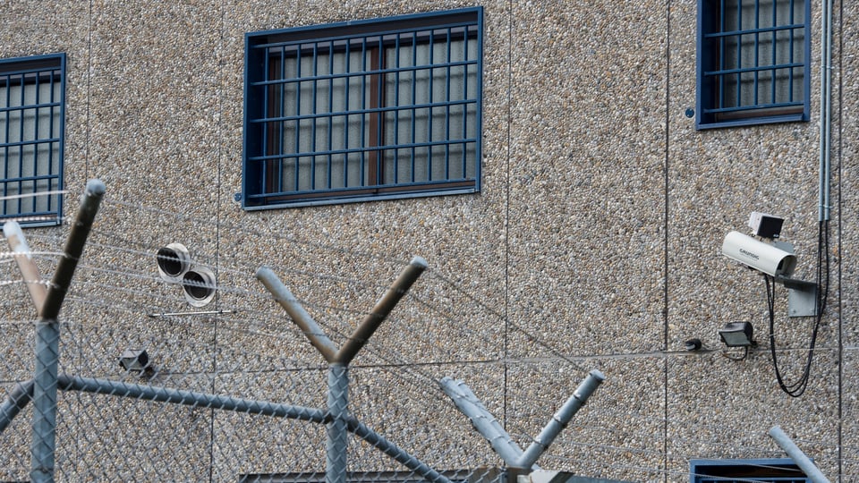 Fassade eines Gefängnisses mit Stacheldraht und vergitterten Fenstern.