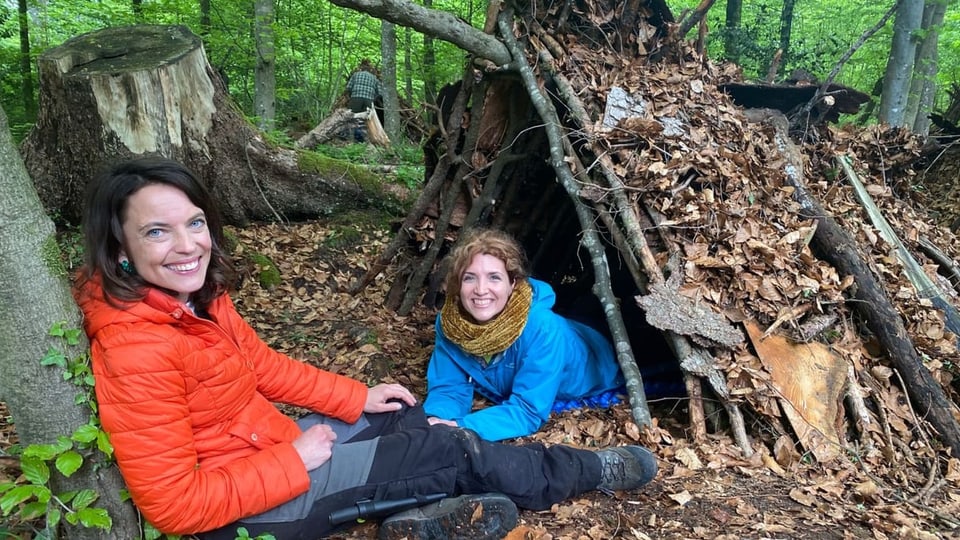 Zwei Frauen bei einer Hütte aus Ästen und Laub.