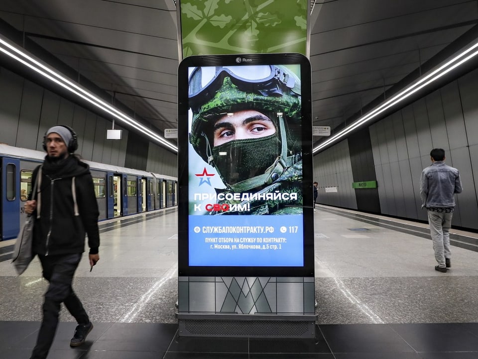Blick auf einen Bahnsteig der Moskauer Metro. Auf einem Plakat ist ein Soldat zu sehen.