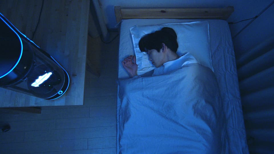 Ein junger Mann schläft auf seinem Bett, neben ihm ist ein Hologramm unter einer Glaskuppel