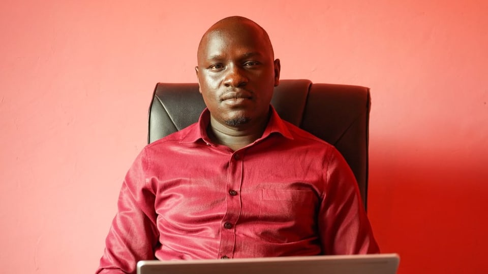 Samuel Okulony sitzt vor seinem aufgeklappten Laptop in einem roten Hemd. 