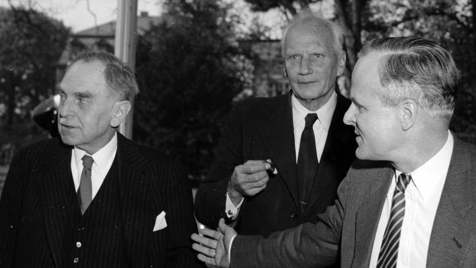 Otto Hahn, Walter Gerlach und Carl Friedrich, alle in Anzug und Krawatte, eilig an der Kamera vorbeigehend.