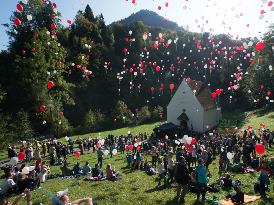 Viele Leute auf einer Wiese mit einer kleinen Kapelle - viele rote und weisse Ballone fliegen in die Luft.