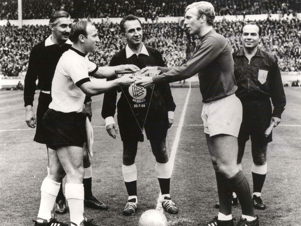 Der Schweizer Schiedsrichter Gottfried Dienst sieht zu, wie die beiden Captains Uwe Seeler und Bobby Moore Wimpel austauschen. Hinten links steht Assistent Tafik Bachramow.
