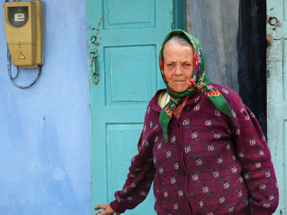 Eine alte Frau mit Kopftuch steht vor ihrem Haus.