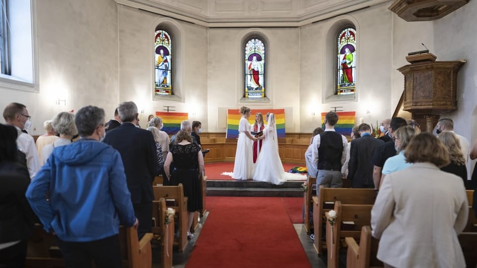 zwei Frauen im weissen Kleid geben sich die Hand, stehen vor Altar. Davor Publikum in Reihen, dahinter Regenbogenflaggen