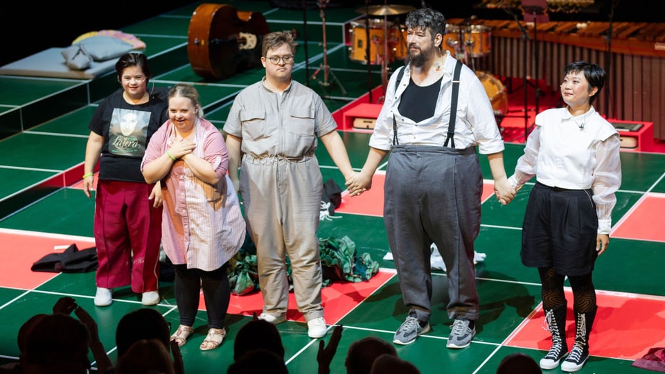 Fünf Personen halten sich an den Händen, sie stehen auf einer rot-grünen Bühne, im Hintergrund Instrumente.