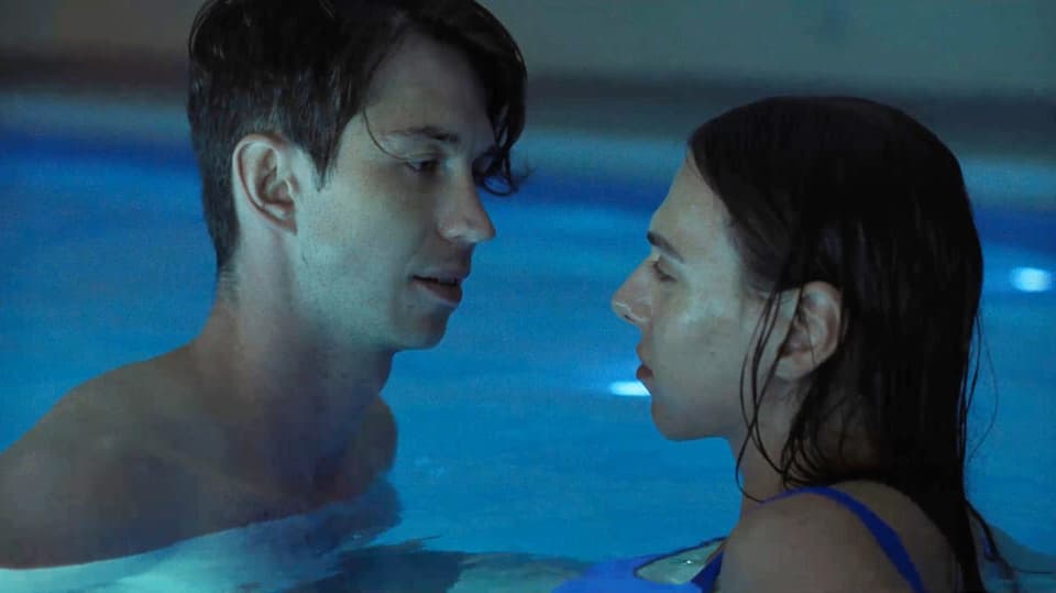 Eine junge Frau und ein junger Mann stehen in einem Swimmingpool und schauen sich in die Augen.