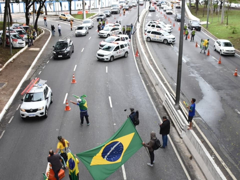 Auf einer Strasse stehen Menschen mit brasilanischen Flaggen und halten Autos an.