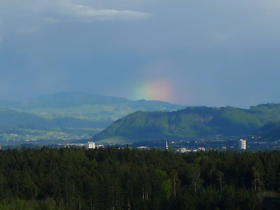 Regenbogensegment über einem Berg, der Himmel ist dunkel teils blinzelt auch die Sonne durch.