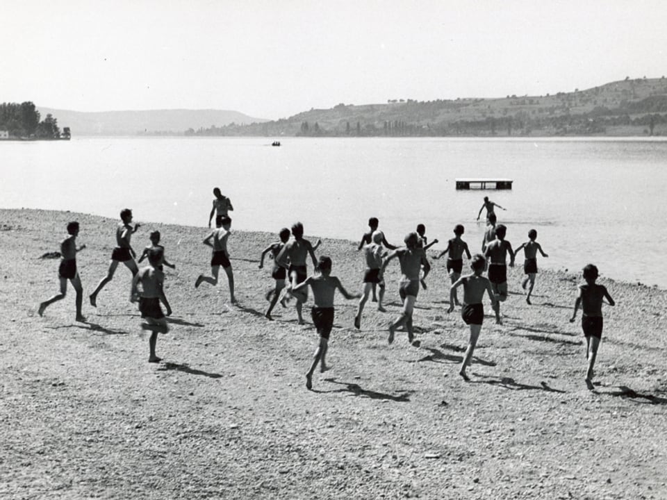 Schwarz-Weiss-Bild: Eine Horde von Kindern läuft in Badehosen auf einen See zu.