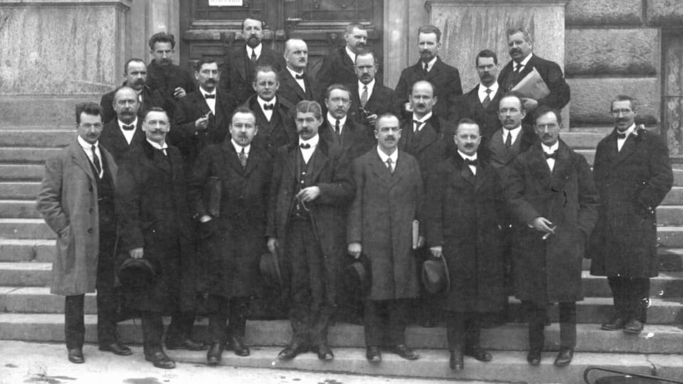 Männer stehen zusammen für ein Gruppenfoto. Alle in schwarzen oder grauen Anzügen.