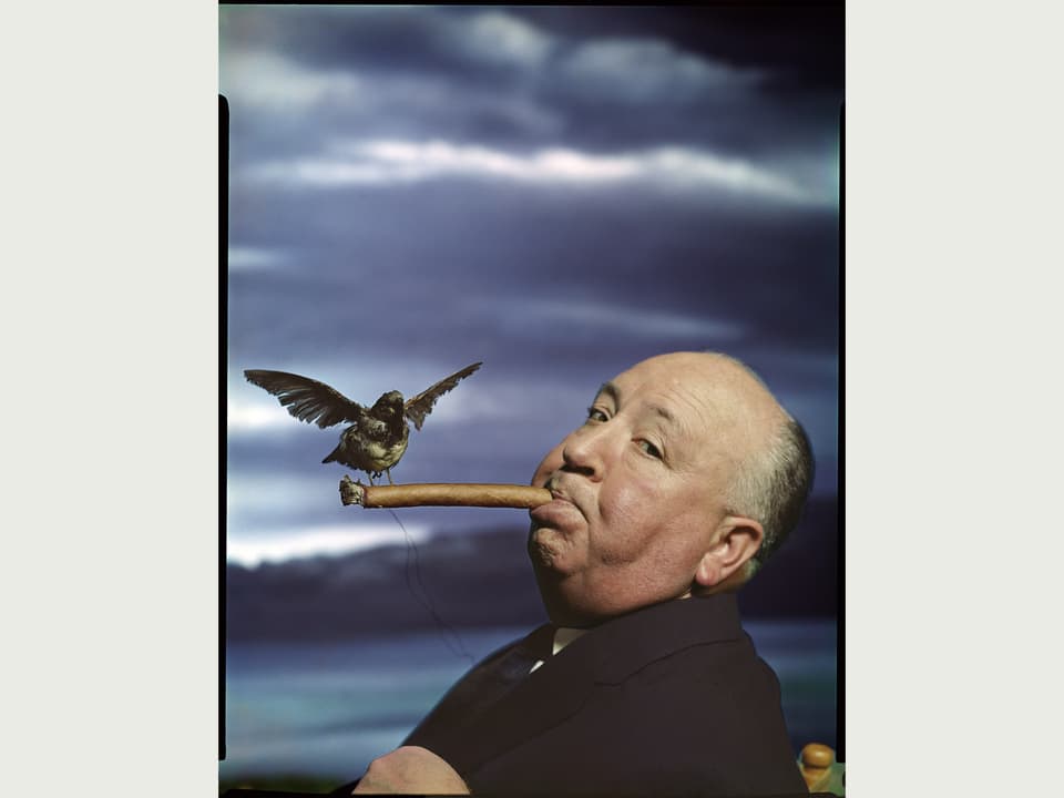 Alfred Hitchcock mit einer Zigarre im Mund, auf der Zigarrenspitze sitzt ein Vogel. Im Hintergrund düstere dunkle Wolken.