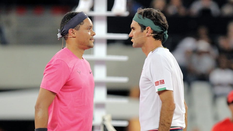 Trevor Noah und Roger Federer stehen auf einem Tennisplatz und blicken sich an. 