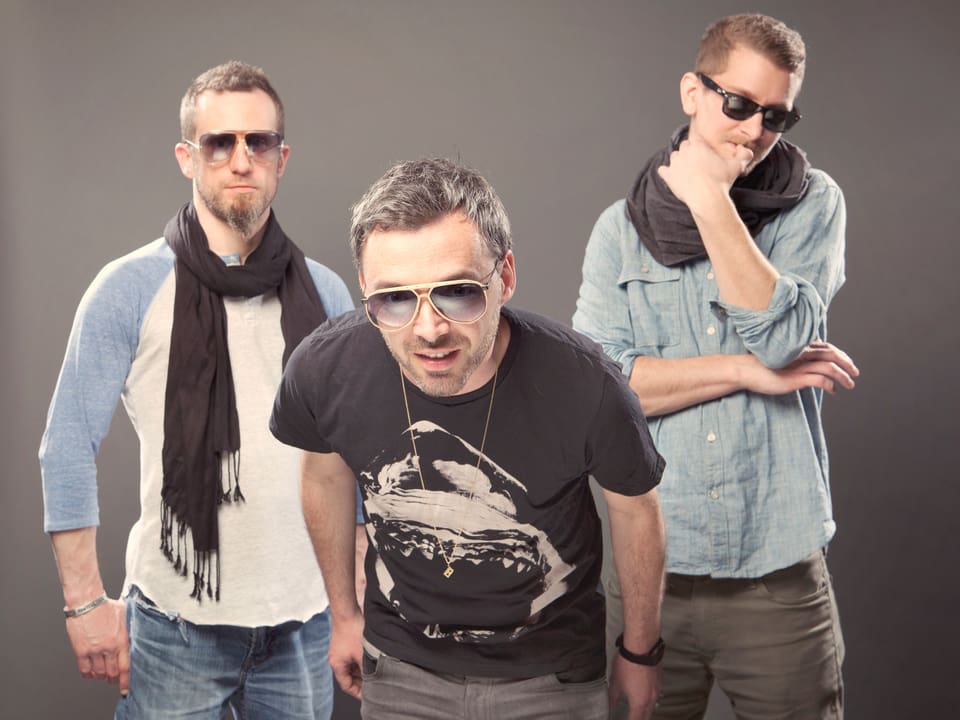 Drei Männer, alle mit Sonnenbrille, stehen vor grauem Hintergrund.