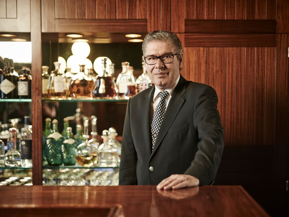 Ein Mann mit Brille in Anzug steht hinter einem dunkelbraunen Bar-Tresen.