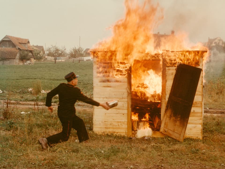 Ein uniformierter Mann vor einem brennenden Häuschen. 