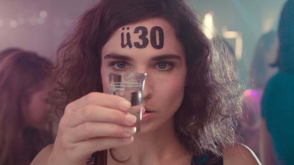 Eine Frau hat ein auf der Stirn "ü30" geschrieben und hält ein Glas Schnaps in der Hand.